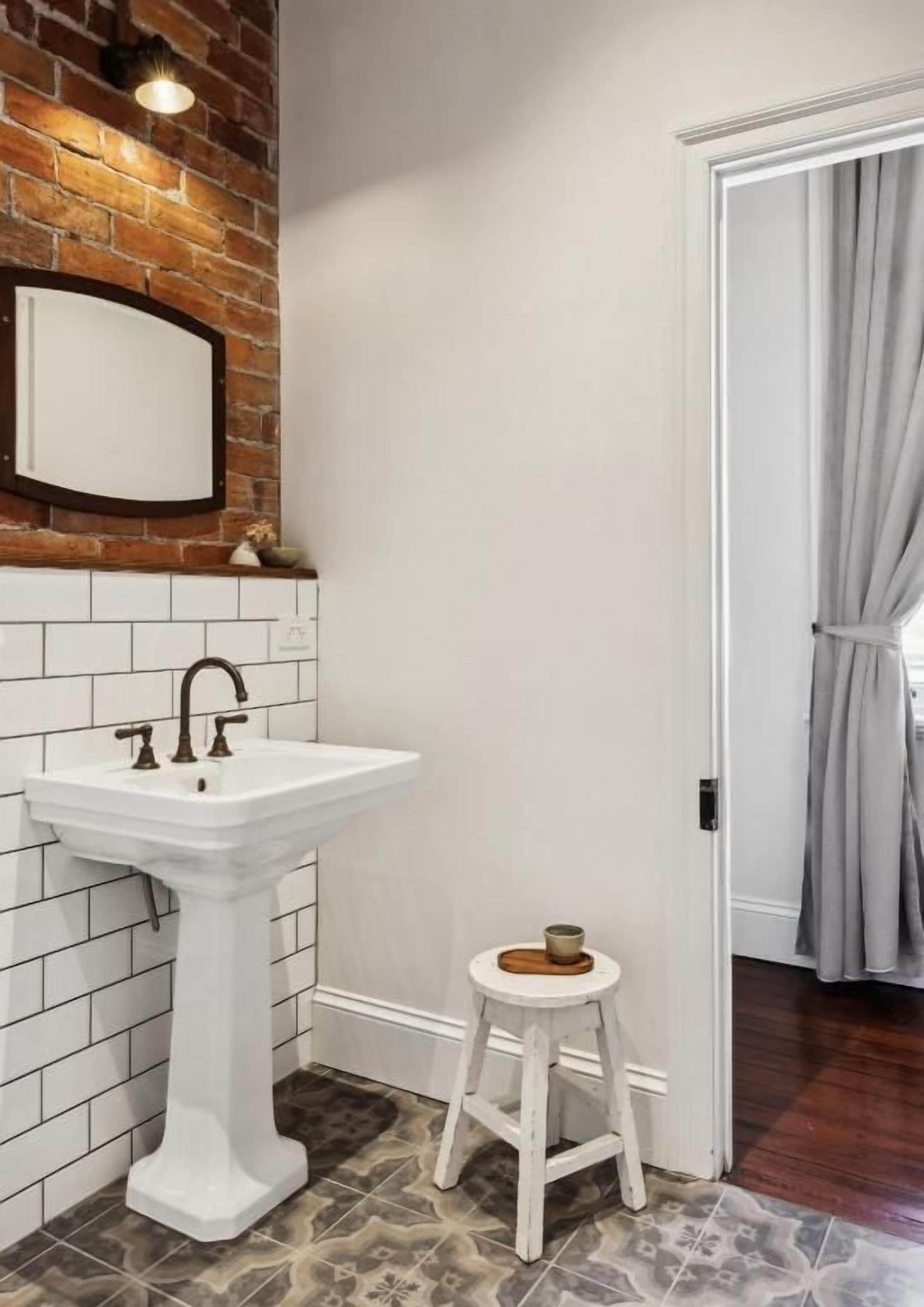 Une salle de bain avec un lavabo sur colonne blanc, un petit tabouret avec un pot, et un miroir accroché à un mur de briques.