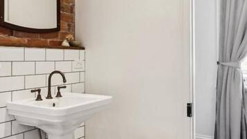 Une salle de bain avec un lavabo sur colonne blanc, un petit tabouret avec un pot, et un miroir accroché à un mur de briques.