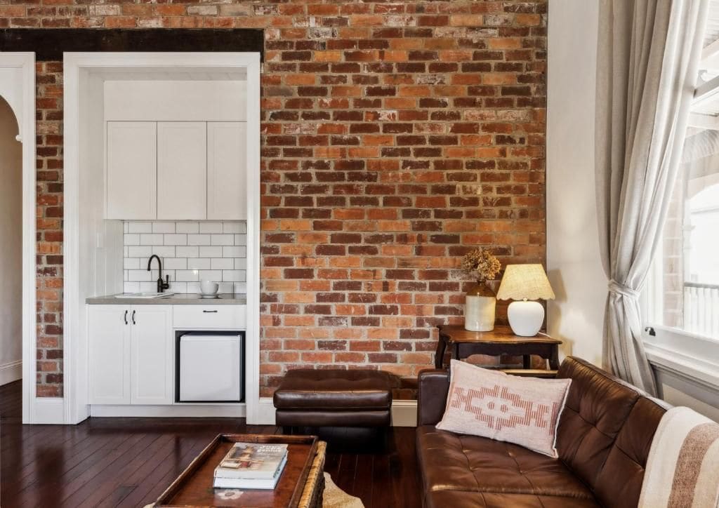 A living room with a brick wall, a brown leather sofa, a white cabinet, and a small table with a lamp.