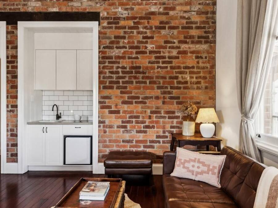 Una sala de estar con pared de ladrillo, un sofá de cuero marrón, un armario blanco y una mesita con lámpara.