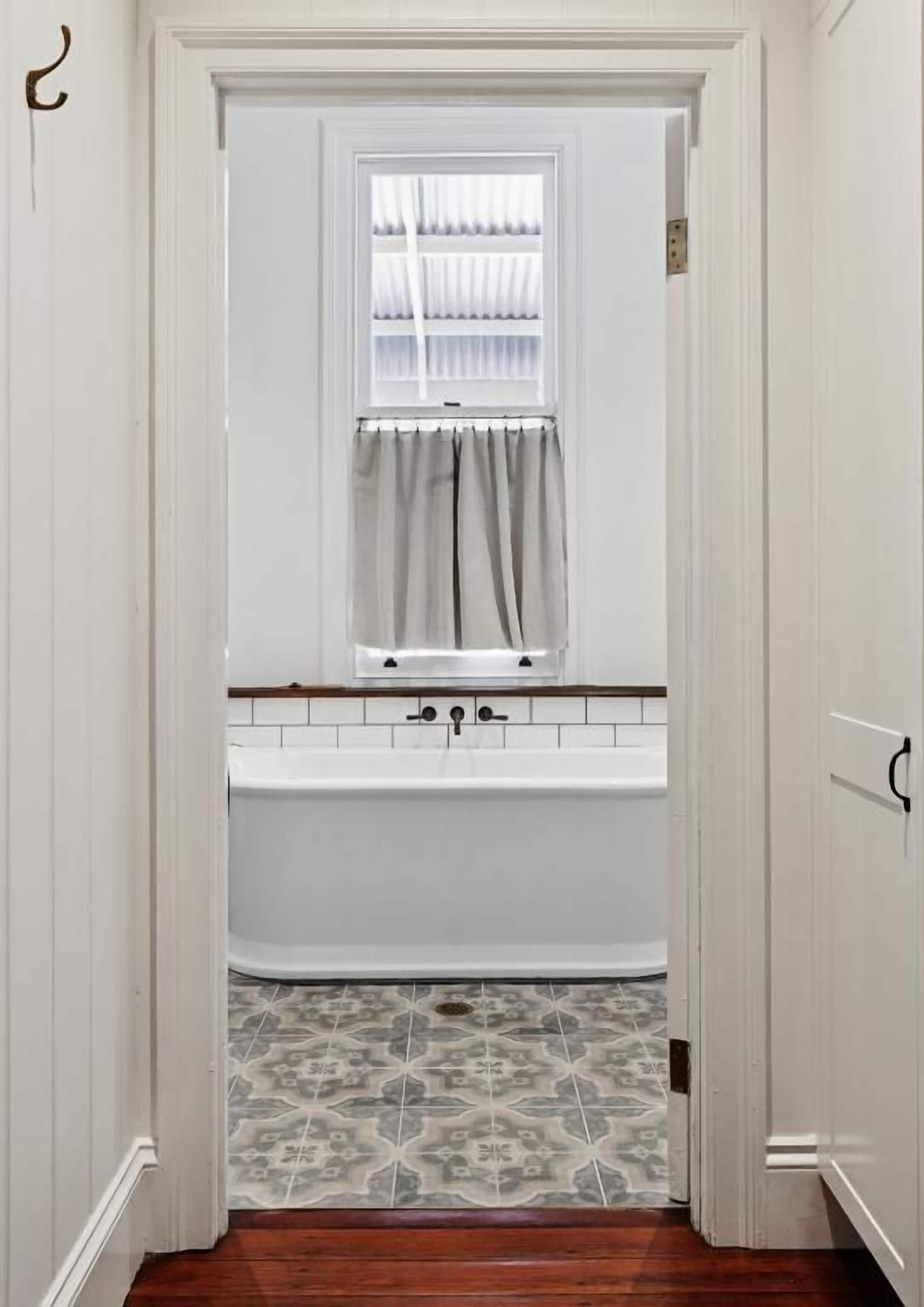 Een badkamer met een witte badkuip, een raam met gordijnen en vloertegels met een patroon.