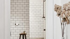 Casa de banho com azulejos brancos tipo metrô, chão estampado, um banquinho pequeno e um vaso com flores secas.