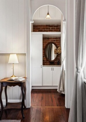 A hallway with a wooden floor, white walls, a round mirror, a table with a lamp, and a hanging light fixture.