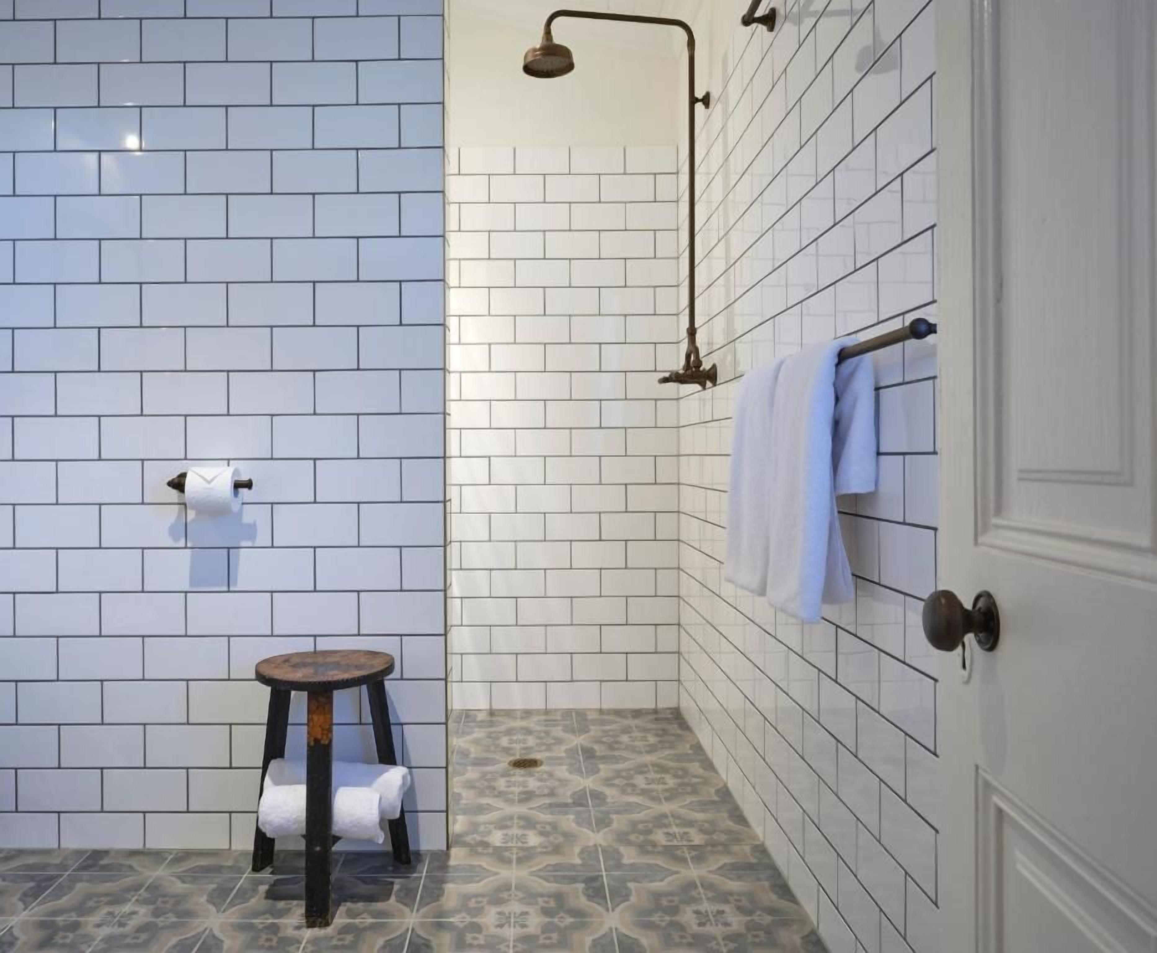 A bathroom with white tiled walls, a shower with a handheld showerhead, a towel rack with a blue towel, and a small stool with a towel.
