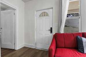 A room with a white door, a red sofa, and a window with curtains. - Cozy Detroit House | 3BR Sleeps 6 Near Downtown (Detroit)