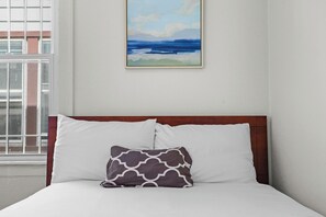 Un lit avec une literie blanche et un oreiller à motifs, un tableau représentant un paysage et une fenêtre donnant sur des bâtiments.
