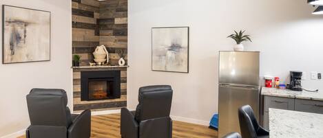 A modern living room with a fireplace, two black leather chairs, a kitchen area with a refrigerator, and abstract art on the walls.