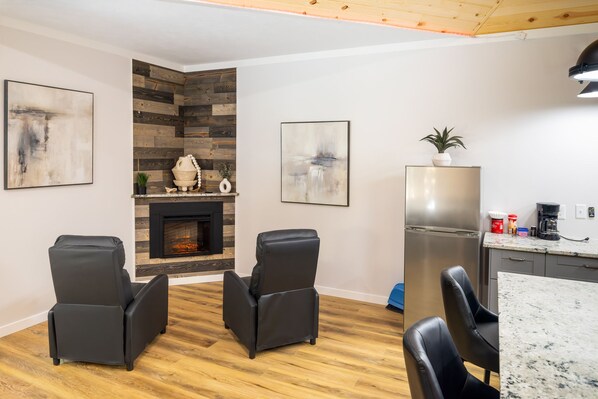 A modern living room with a fireplace, two black leather chairs, a kitchen area with a refrigerator, and abstract art on the walls.