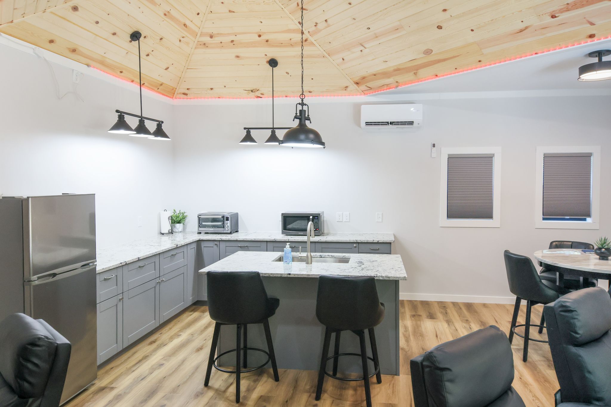 A modern kitchen with a central island, grey cabinets, stainless steel appliances, pendant lighting, and a wooden ceiling.