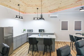 Eine moderne Küche mit Kochinsel, grauen Schränken, Edelstahlgeräten, Pendelleuchten und einer Holzbalkendecke.