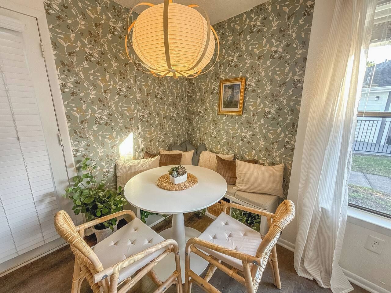 Quarto com mesa redonda, cadeiras de vime, um sofá e uma janela grande com cortinas.