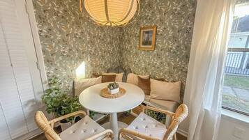 Quarto com mesa redonda, cadeiras de vime, um sofá e uma janela grande com cortinas.