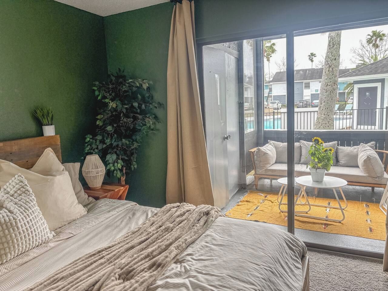 A bedroom with a bed, a potted plant, and a view of a balcony with outdoor furniture.