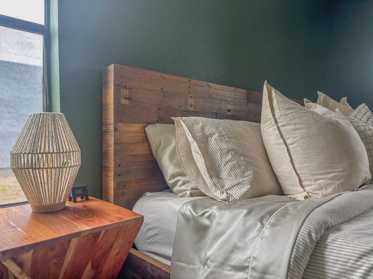 Un dormitorio con cabecera de madera, cama con muchos almohadones y mesita de noche de madera con una lámpara.