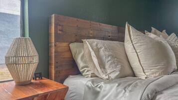 Un dormitorio con cabecera de madera, cama con muchos almohadones y mesita de noche de madera con una lámpara.