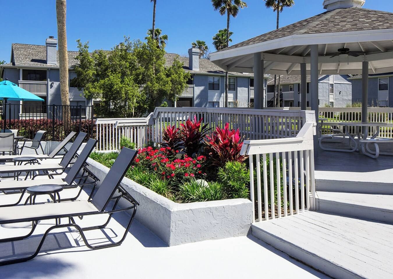 Un espace aménagé au bord de la piscine, avec des chaises longues, un coin salon abrité et un jardin agrémenté de fleurs rouges.