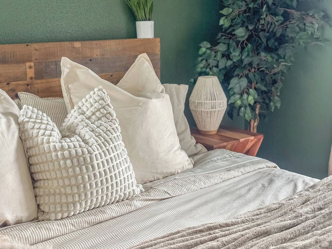 Ein Bett mit gestreifter Bettwäsche, mehreren Kissen, einem hölzernen Kopfteil, einem Nachttisch mit Lampe und einer Topfpflanze.