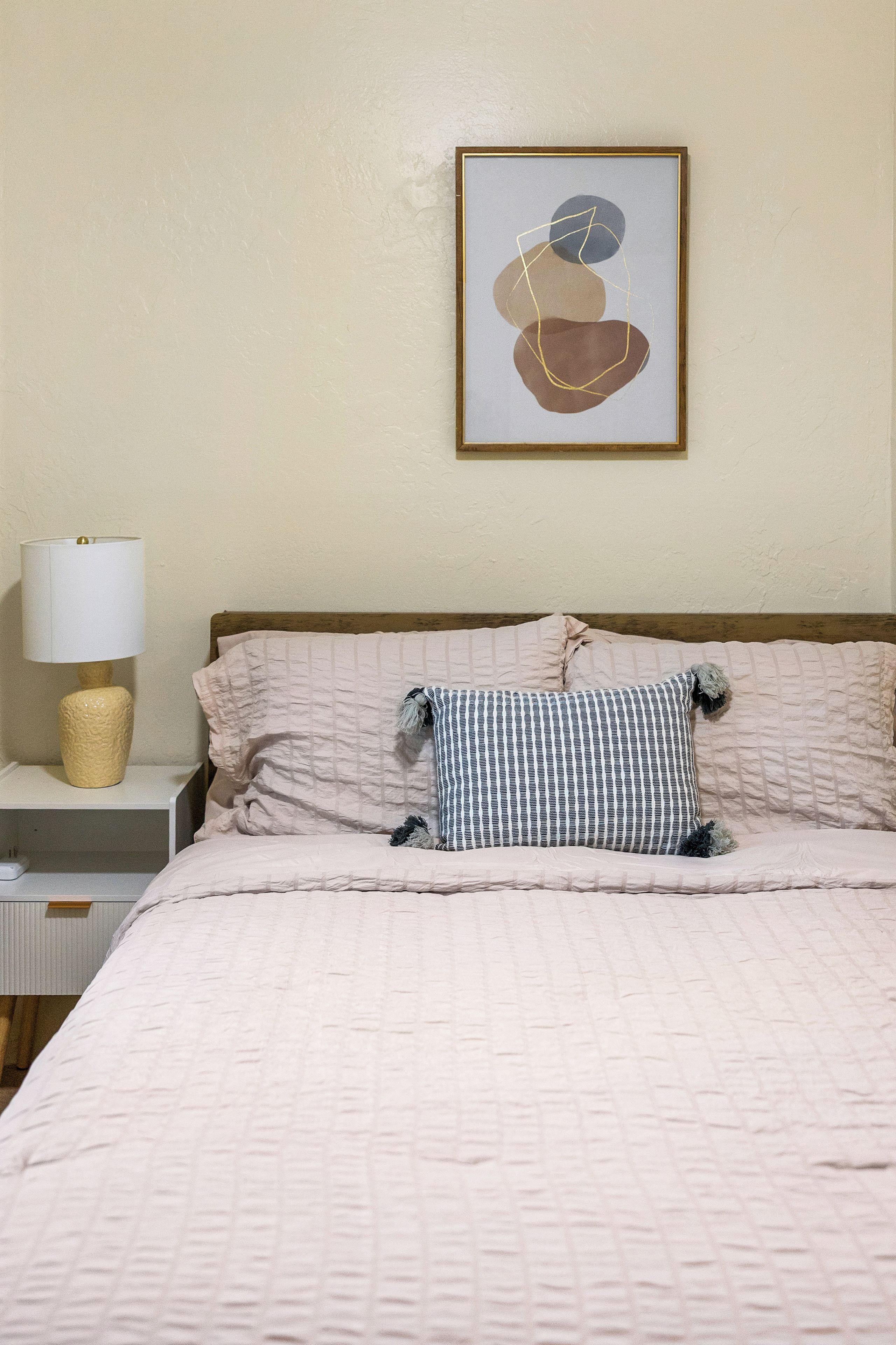 Een slaapkamer met een bed, kussens, een lamp en een lijst met een abstract schilderij aan de muur.