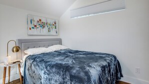 Ein Schlafzimmer mit einem Bett, einem Nachttisch, einer Lampe, einem Bild und einem Fenster mit Jalousien.