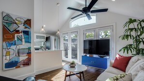 Ein modernes Wohnzimmer mit Deckenventilator, einem Essbereich und einer dahinterliegenden Küche, sowie einem großen abstrakten Gemälde an der Wand.