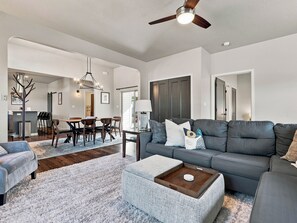 A spacious living room with a grey sofa, a coffee table, and a dining area with chairs and a table. - GJ Getaway - Fire Pit + Bocce + Private Yard! (Grand Junction)