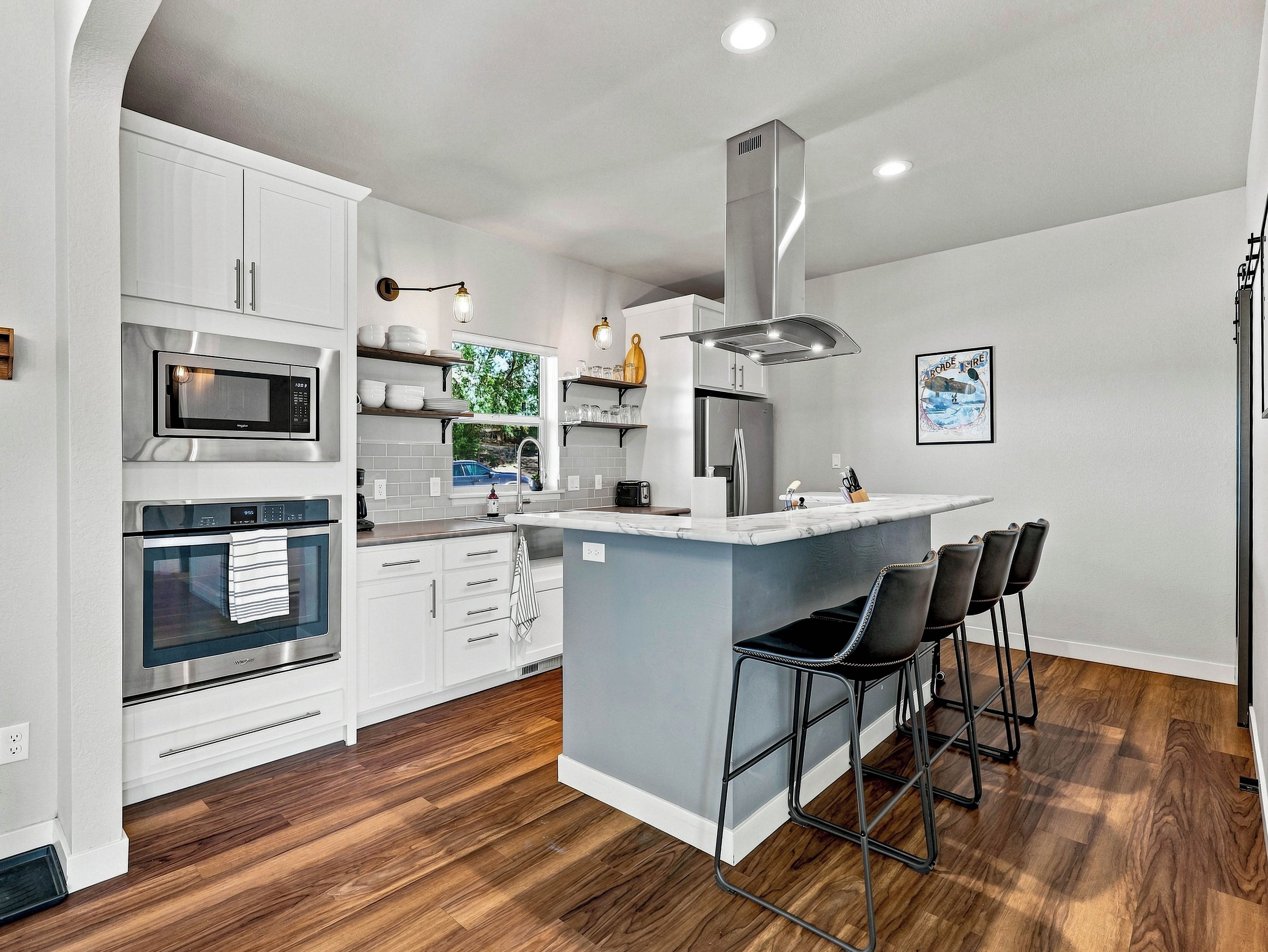A modern kitchen with white cabinetry, stainless steel appliances, a central island with bar stools, and a large window.