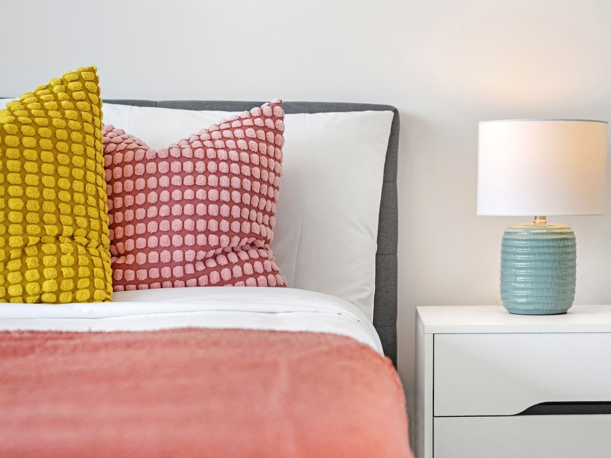 Un lit recouvert d’une couverture blanche et rose, deux coussins à motifs, une table de chevet avec une lampe et une tête de lit grise.