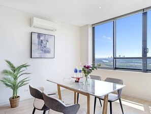 A modern room with a large window, a table with chairs, a potted plant, and a framed picture on the wall. - High-Rise 2BR Retreat with Water Views (Wentworth Point)