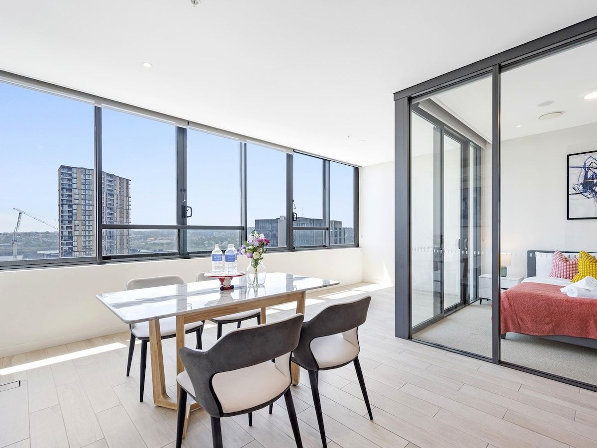 Een moderne hotelkamer met een groot raam, een zithoek met een tafel en stoelen, en een bed met een rode deken.