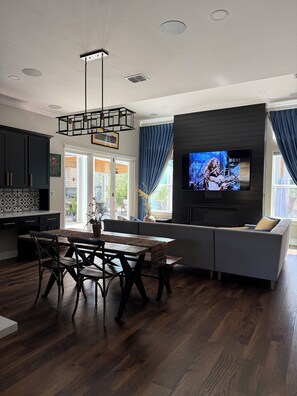 A modern living room with a dining area, a flat-screen TV mounted on a wall, and a large window with curtains. - Charming 3-bedroom house in Canyon Lake with close access to 3 boat ramps. (Canyon Lake)