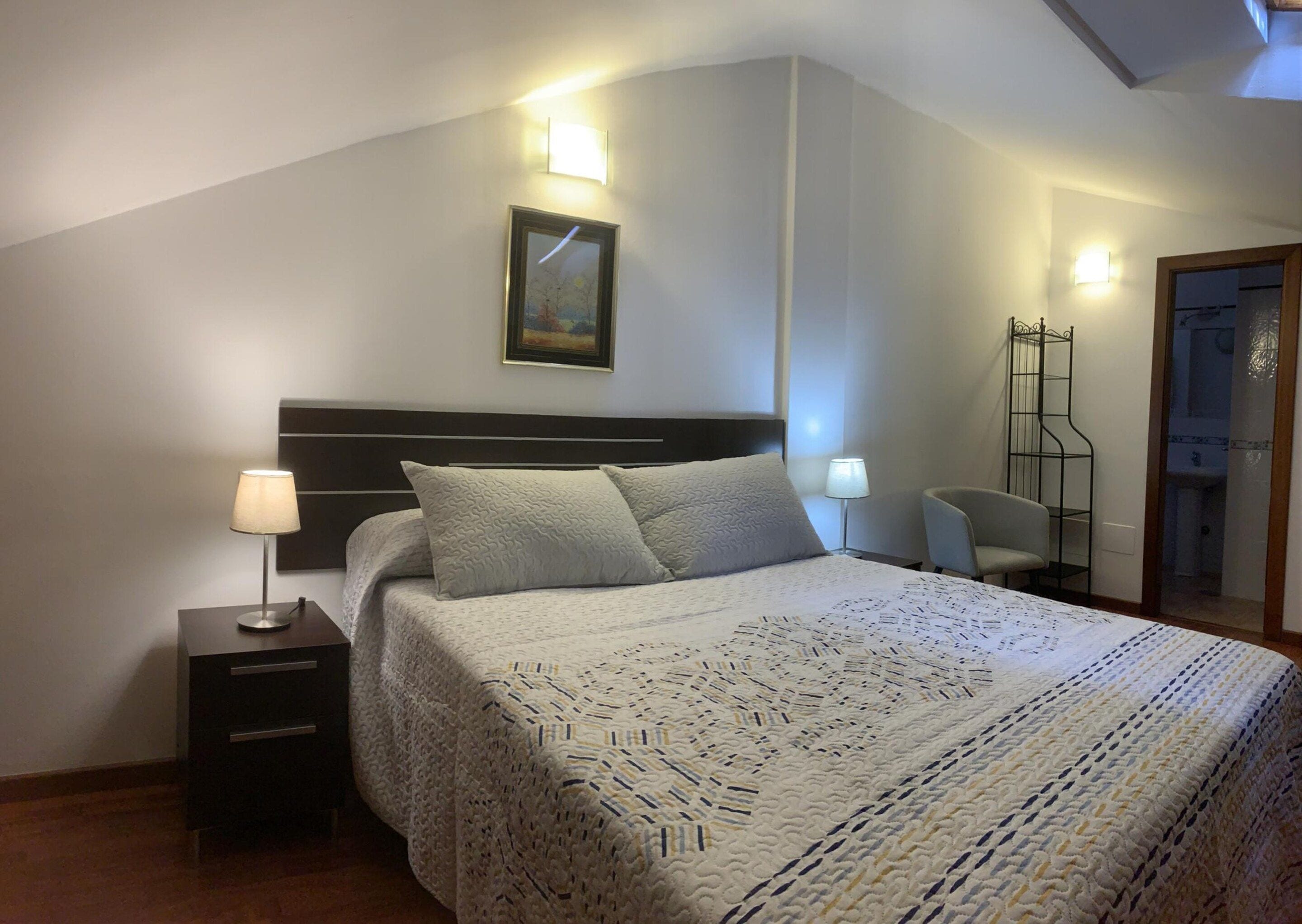 A bedroom with a bed, bedside lamps, a nightstand, a chair, and a painting on the wall.