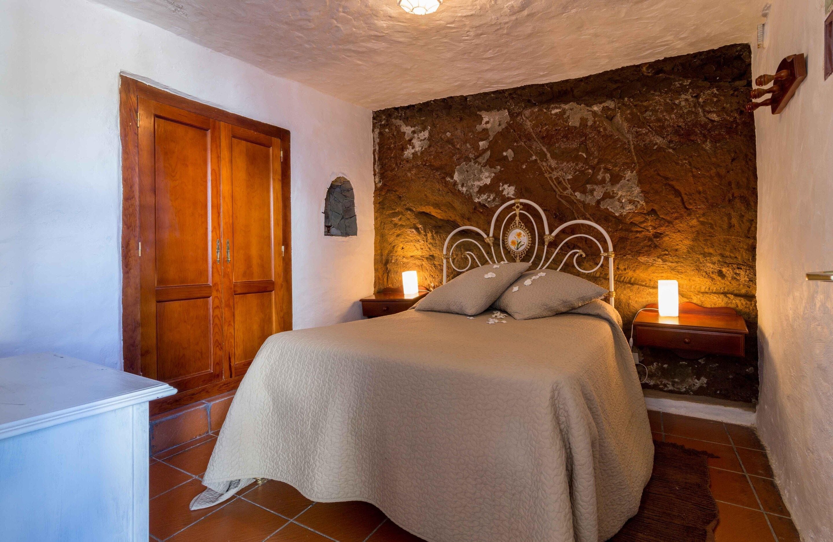 Quarto com cama, guarda-roupa de madeira, parede de pedra e duas luminárias de cabeceira.