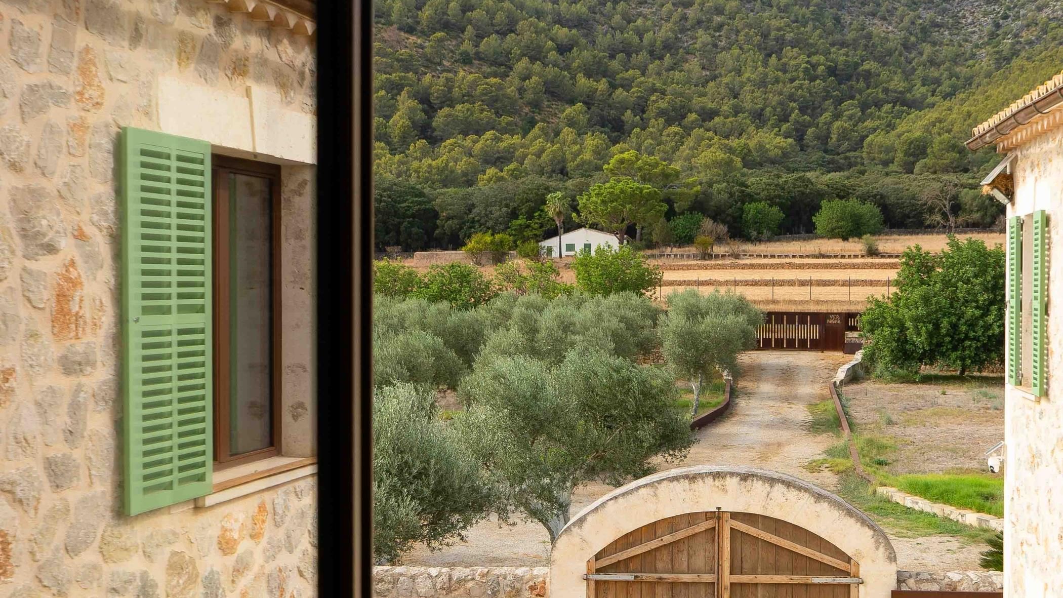 Un bâtiment en pierre doté d’une fenêtre et de volets, un chemin menant à un portail et une vue sur une colline boisée.