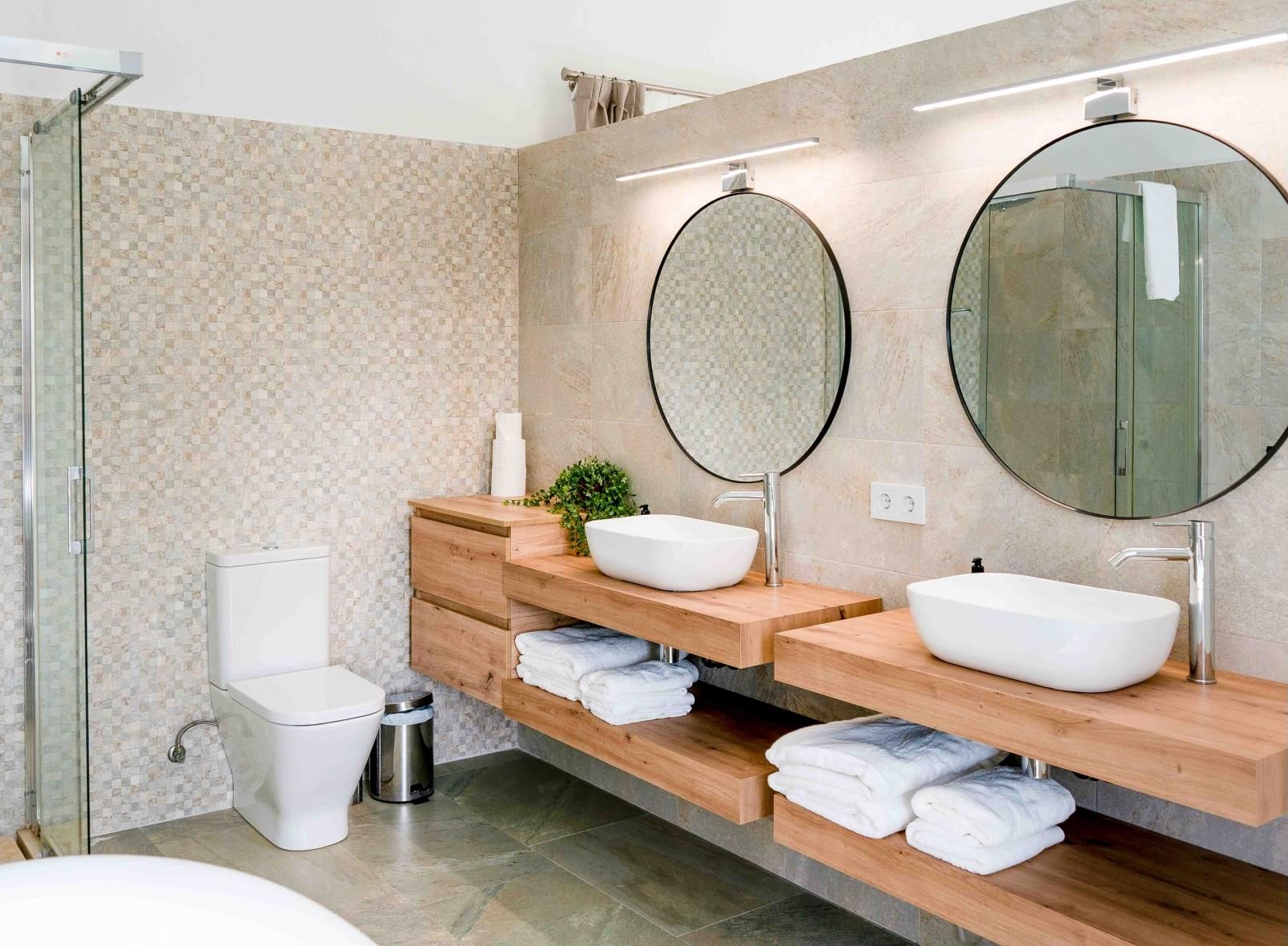 A modern bathroom with a glass shower enclosure, a toilet, a bidet, and two sinks with round mirrors.