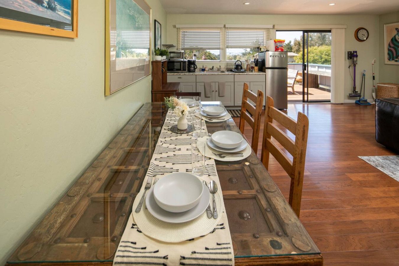 A dining area with a long table set for four, a kitchen in the background, and a view of the outdoors.