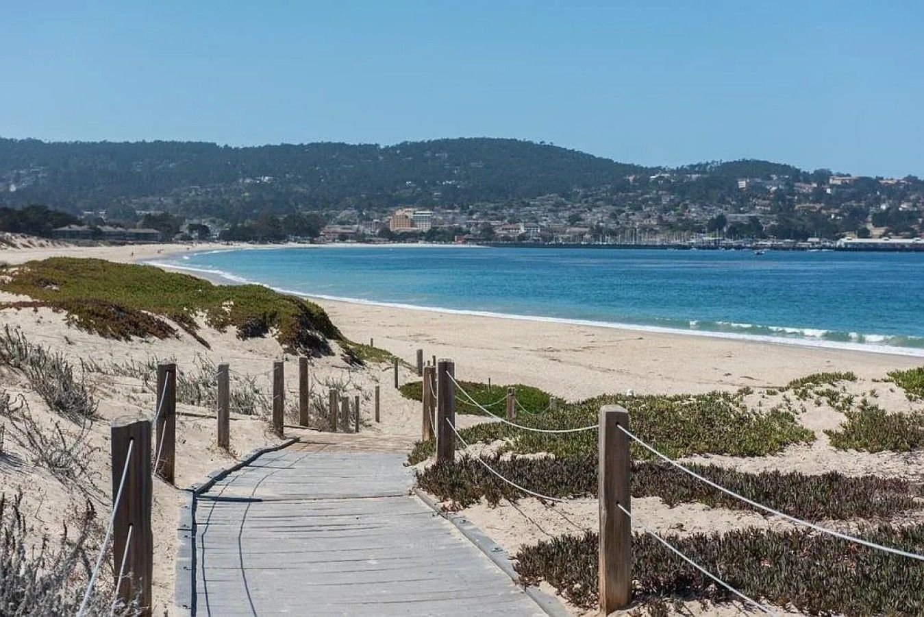 A coastal walkway leading to a beach with a clear view of the ocean and a distant town.