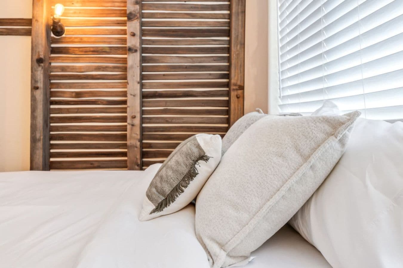 Una cama con sábanas blancas y dos almohadas, pared de listones de madera y lámpara fijada a la pared.