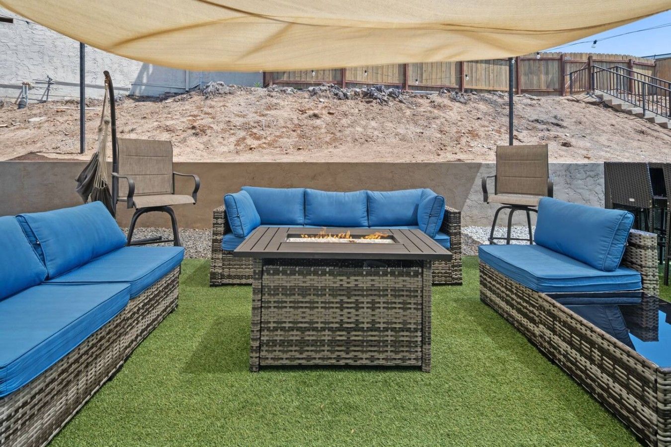 An outdoor seating area with wicker furniture and blue cushions, a fire pit, and a backdrop of a sandy hill.