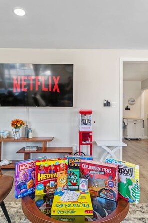 Un salon avec un téléviseur à écran plat affichant le logo de Netflix, une machine à café et une variété de jeux de société disposés sur une table basse.
