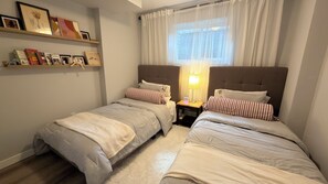 A bedroom with two single beds, a bedside table with a lamp, a window with sheer curtains, and a shelf with framed pictures and books.
