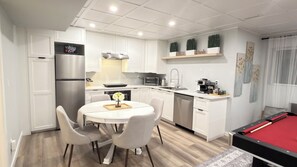 Cocina compacta con armarios blancos, una mesa de comedor con sillas, una cafetera y una mesa de billar.