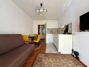 A modern living room with a sofa, a dining table with yellow chairs, a kitchen area with white cabinets, and a ceiling light fixture. - Kościuszki Central Apartment #7 (Wrocław)
