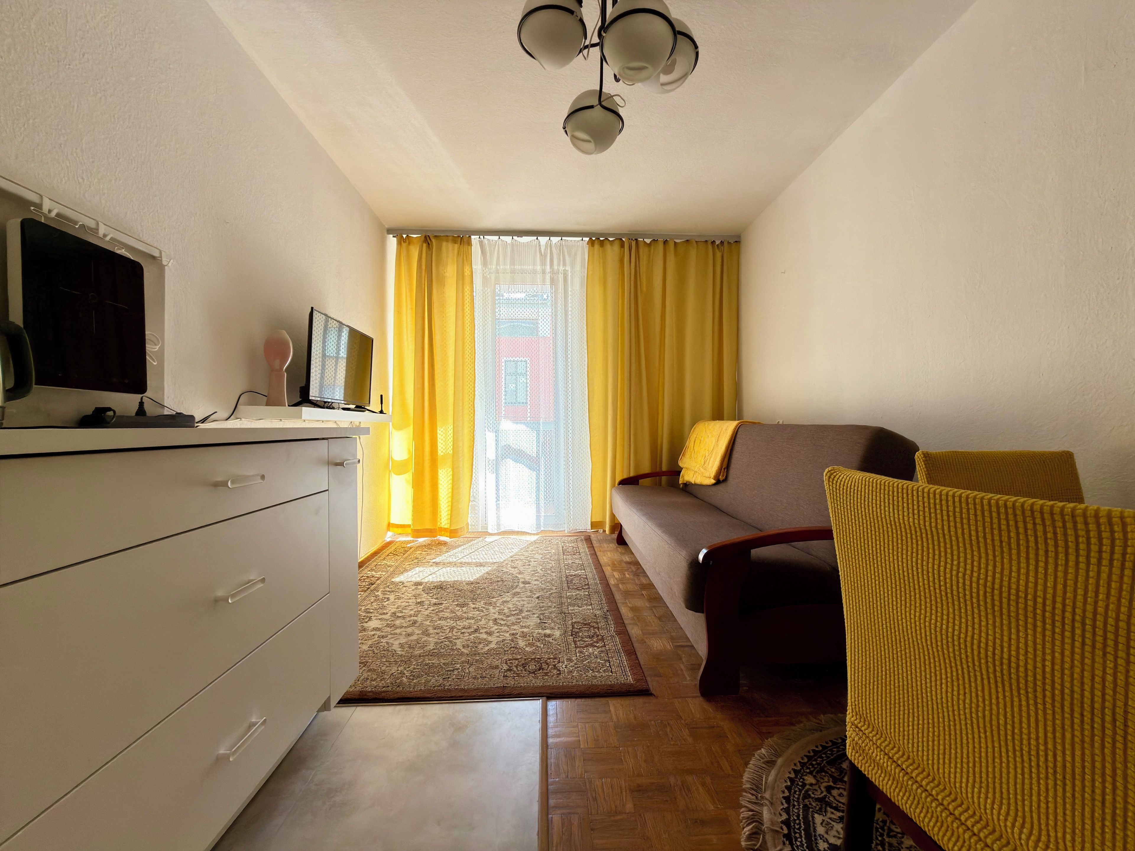 Une pièce comprenant un canapé, un téléviseur posé sur un meuble, un bureau avec des tiroirs, un tapis à motifs et une fenêtre avec des rideaux jaunes.