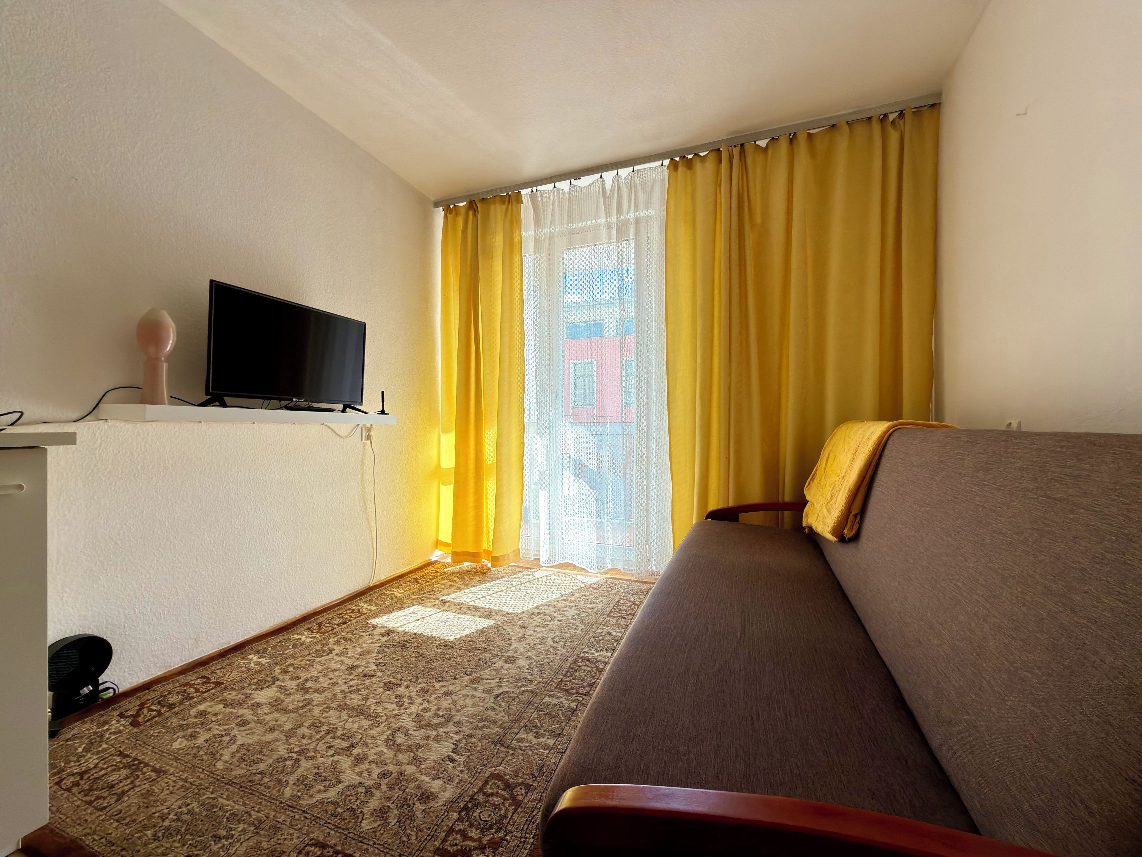 A hotel room with a sofa, a television on a stand, a patterned carpet, and a window with yellow curtains.