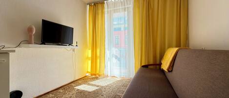 A hotel room with a sofa, a television on a stand, a patterned carpet, and a window with yellow curtains.