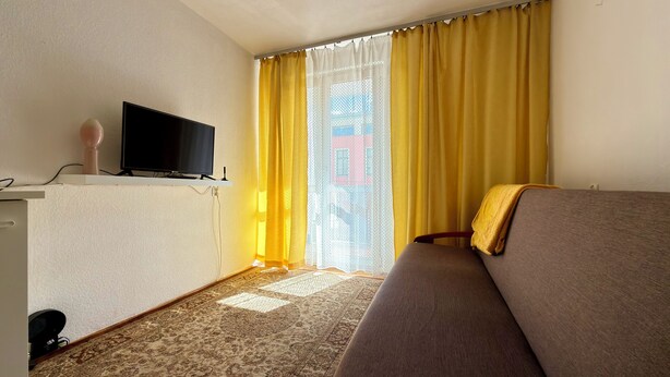 Ein Hotelzimmer mit einem Flachbildfernseher auf einem Gestell, einem Sofa, einem gemusterten Teppich und einem Fenster mit gelben Vorhängen.