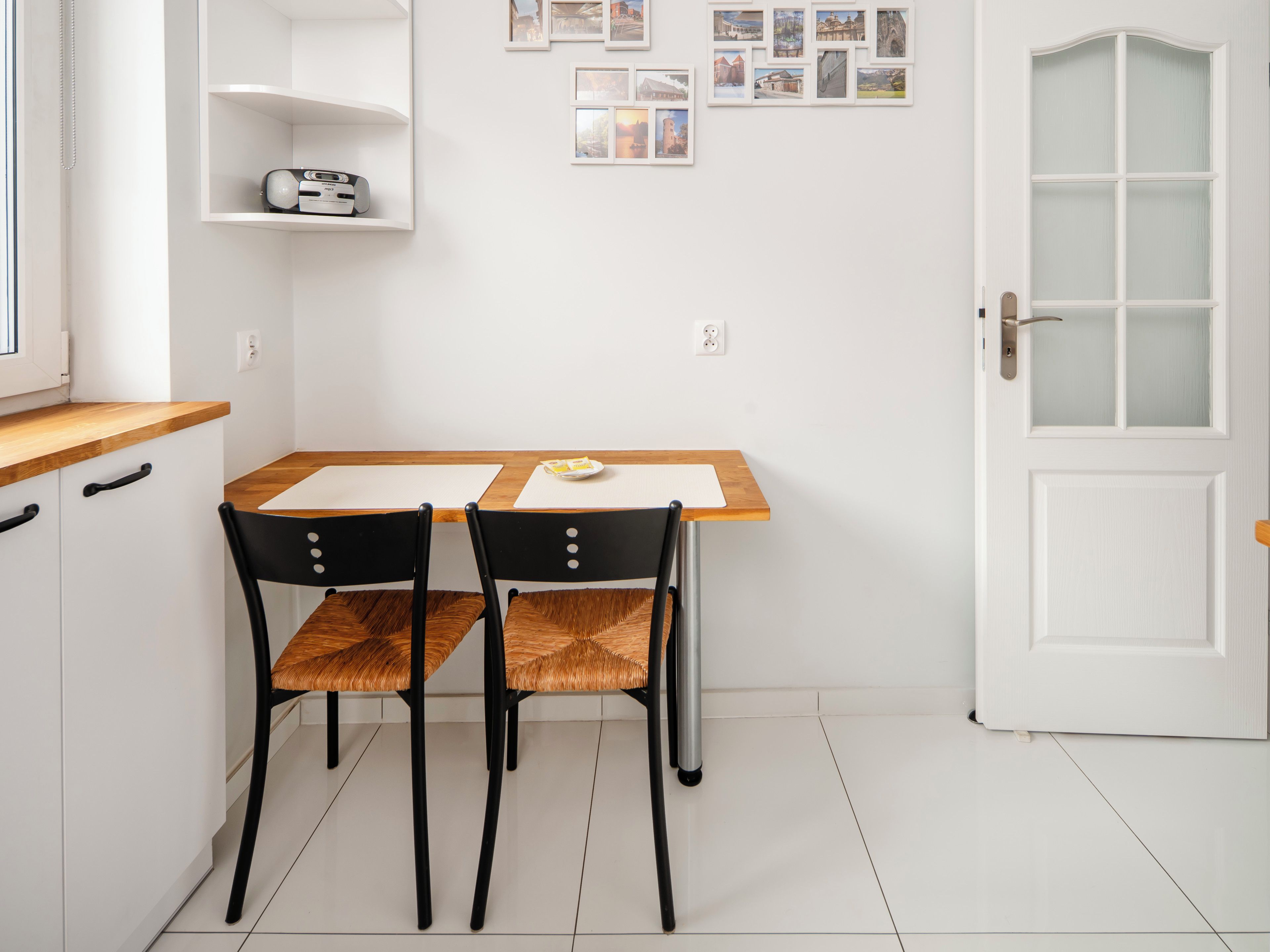 Une petite pièce propre, avec une table en bois et deux chaises, une porte blanche à panneaux de verre et une étagère fixée au mur sur laquelle se trouve une radio.