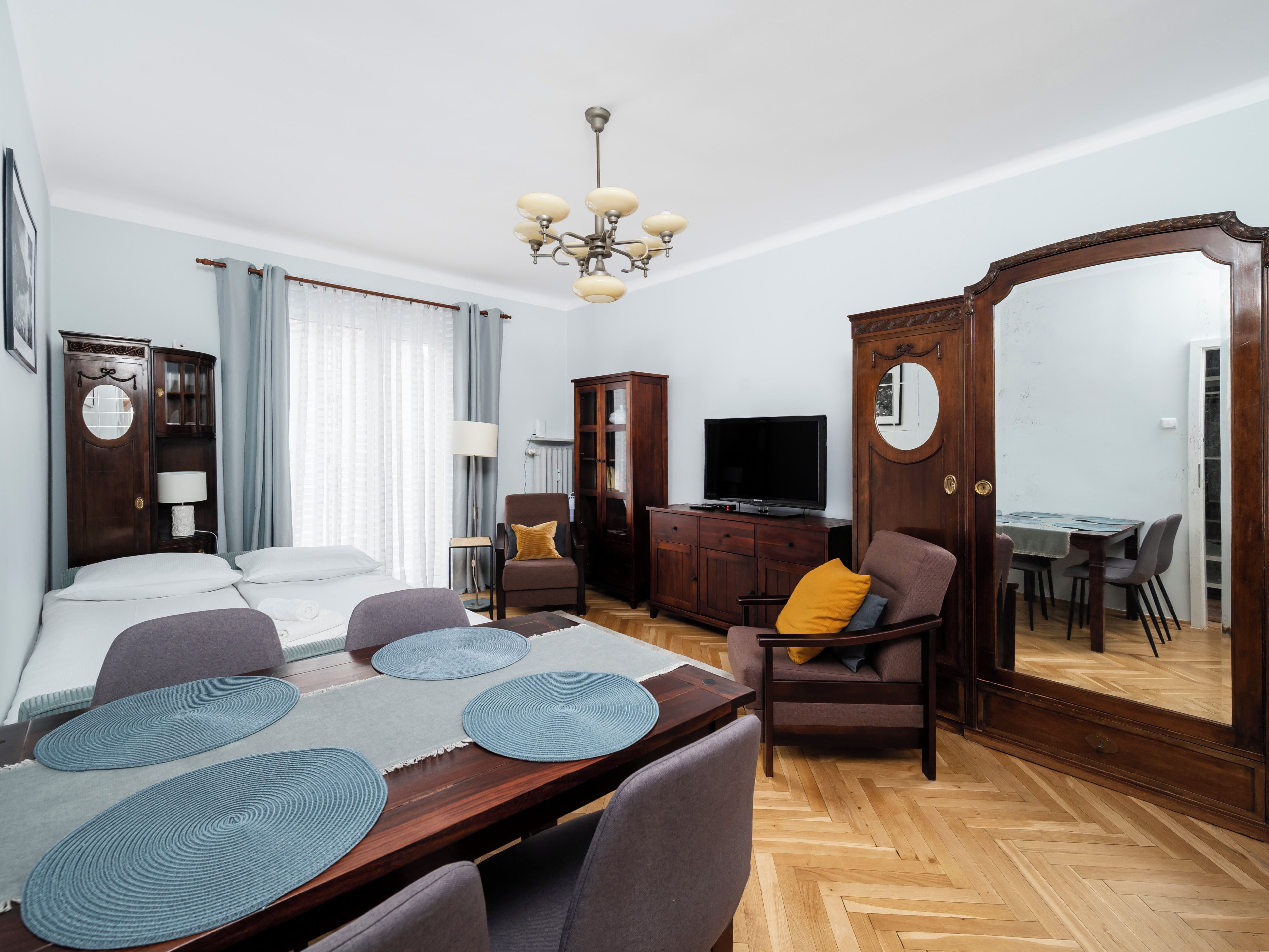 Une chambre d’hôtel comprenant un lit, une table à manger avec des sets de table, un téléviseur, une armoire et un grand miroir.