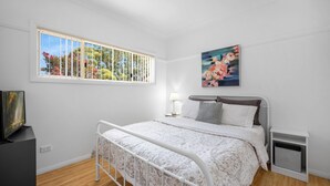 Een slaapkamer met een bed, een raam met luxaflex, een bedlampje, een schilderij en een tv op een standaard.