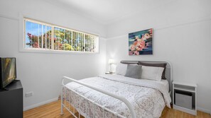 Un dormitorio con cama, ventana con persianas, lámpara de noche, un cuadro y televisor sobre un mueble.